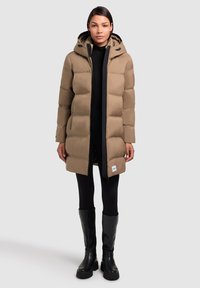 Abrigo acolchado beige con capucha, que presenta un cierre frontal, bolsillos laterales y un parche con logo. Se lleva sobre leggings negros y botas altas negras.