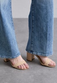 Sandali aperti ai piedi di colore beige con due fasce larghe, tacco a blocco e texture liscia, abbinati a jeans a gamba larga di colore blu chiaro.