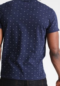 T-shirt en coton bleu marine avec un petit motif d'ancre, manches courtes et coupe ajustée. Texture lisse avec un col rond.