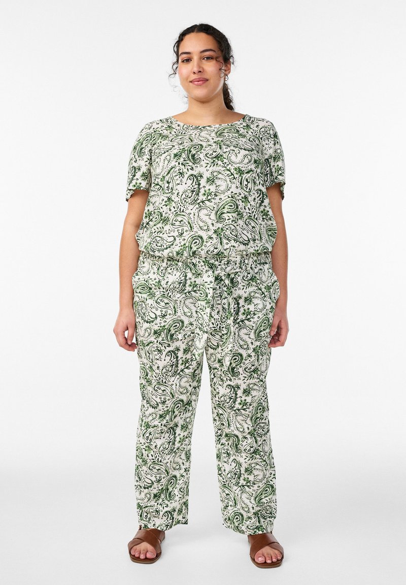 Donna con top a maniche corte bianco e verde con stampa paisley e pantaloni coordinati, in piedi con le mani rilassate, sandali marroni visibili.