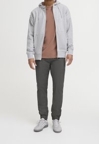 Grijze hoodie met rits, taupe t-shirt, donkergrijze taps toelopende broek en lichtgrijze sneakers. Voorzien van trekkoorden en omgeslagen enkels.