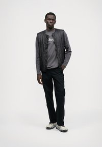 Μαύρο ελαφρύ γιλέκο πάνω από γκρι μακρυμάνικο πουκάμισο, συνδυασμένο με μαύρο παντελόνι και γκρι αθλητικά παπούτσια. Λείο υλικό, απλός σχεδιασμός.