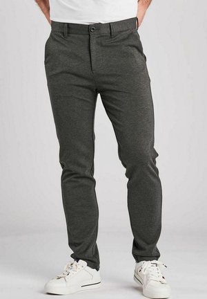 Mand iført mørkegrå slim-fit bukser og hvide sneakers, stående mod en ensfarvet baggrund.