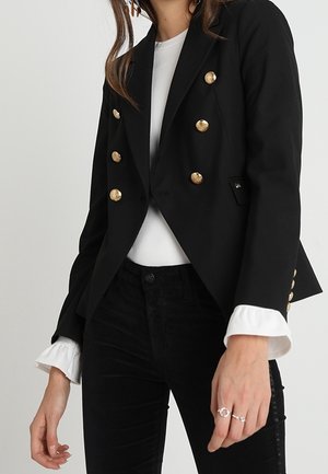 Vrouw draagt een zwarte dubbelgeknoopte blazer met gouden knopen, een witte blouse met ruches aan de manchetten en een zwarte broek, toont hand met ringen.