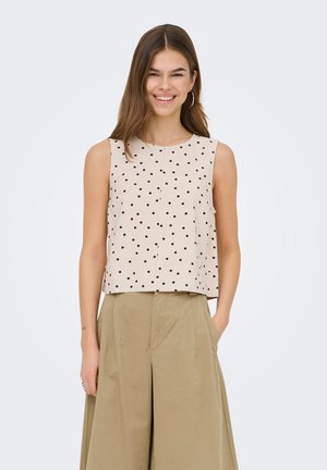 Mujer sonriente que lleva un top beige sin mangas con lunares negros y pantalones anchos de tiro alto color caqui, de pie con una mano en el bolsillo.