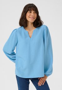 Blouse bleu clair, texturée, avec un col en V et des manches bouffantes. Coupe relaxée avec un ourlet droit, portée avec un jean bleu.