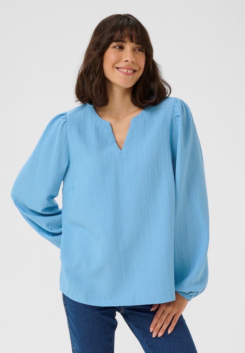 Blouse bleu clair, texturée, avec un col en V et des manches bouffantes. Coupe relaxée avec un ourlet droit, portée avec un jean bleu.