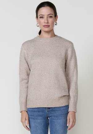 Suéter de punto color beige claro con cuello redondo, puños acanalados y dobladillo acanalado. Los acentos brillantes crean textura a lo largo de la tela. Combinado con jeans azules.