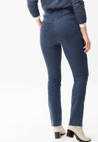 Jean en denim bleu foncé avec une silhouette ajustée, taille haute, deux poches arrière et des coutures contrastées. Associés à des bottines à talons beiges.