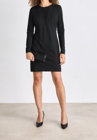 Černé šaty s dlouhým rukávem, výstřihem do V, přiléhavým střihem a měkkou tkaninou. Kombinované s černou strukturovanou psaníčkovou kabelkou a botami s špičatou špičkou.