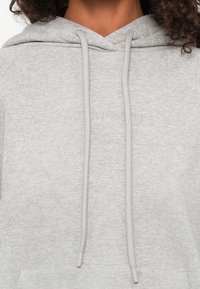 Grijze hoodie van zacht materiaal, met een centrale kangoeroezak en trekkoorden met plastic uiteinden. Subtiel logo in reliëf.