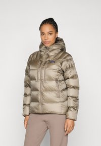 Vrouw in een taupe pufferjack met capuchon, twee verticale borstzakken met rits en een voorrits, staand tegen een effen lichte achtergrond.