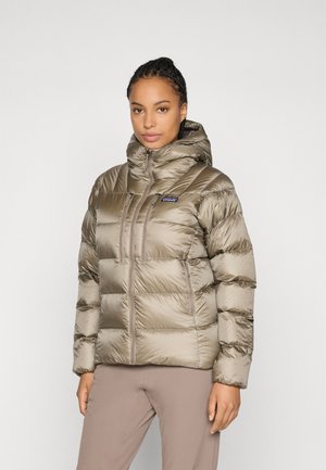 Vrouw in een taupe pufferjack met capuchon, twee verticale borstzakken met rits en een voorrits, staand tegen een effen lichte achtergrond.