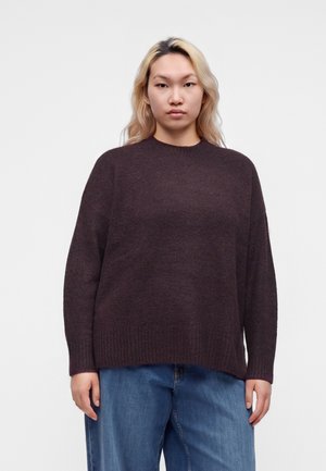 Personne aux longs cheveux blonds portant un pull marron foncé et un jean bleu, debout devant un fond clair uni.