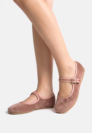 Persona che indossa ballerine Mary Jane in camoscio rosa con fibbie, viste dalla metà del polpaccio in giù su uno sfondo chiaro e uniforme.