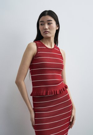 Γυναίκα με μακριά σκούρα μαλλιά που φοράει αμάνικο κόκκινο πλεκτό τοπ και φούστα με λευκές οριζόντιες ρίγες, στέκεται μπροστά σε έναν απλό γκρι τοίχο.