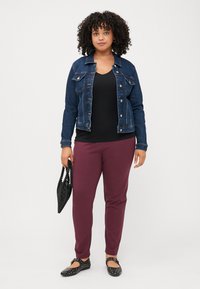 Oseba s kodrastimi lasmi nosi usnjeno jakno čez črn top, bordo hlače in črne balerinke, v roki drži črno torbo. Nevtralna ozadje.