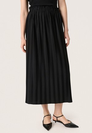 Woman wearing a long black pleated skirt and black pointed-toe low heel shoes with ankle straps, standing on a light background.