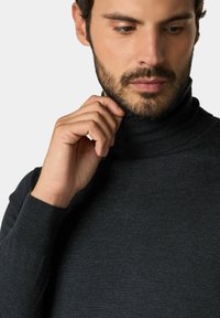 Uomo con la barba che sistema il colletto di un dolcevita grigio scuro, guardando verso il basso su uno sfondo semplice.