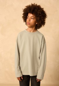 Helehall pikkade varrukatega särk, lõdva lõikega, ümmarguse kaelusega ja soonikkoes varrukate äärtega, kombineeritud tumedate pükstega, neutraalse taustaga.