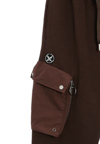 Pantalones cargo marrones con un gran bolsillo lateral con cierre de botón a presión y cremallera, acentuados por un parche negro circular y herrajes de metal.