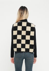 Gilet a motivo a scacchiera in tessuto morbido nero e crema, senza maniche, tagliato all'altezza della vita, indossato sopra una maglietta nera a maniche lunghe.
