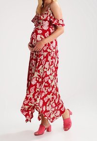 Rode maxi-jurk met bloemenprint en ruches aan de mouwen, voorzien van roze en witte bloemmotieven; gecombineerd met roze sandalen met blokhak.