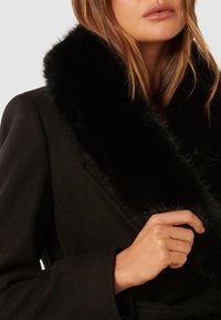 Kobieta w ciemnym płaszczu z grubym czarnym futrzanym kołnierzem, trzyma kołnierz jedną ręką blisko twarzy.