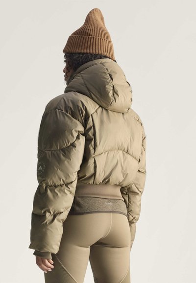 Persona que lleva un gorro marrón de punto, una chaqueta acolchada corta verde oliva y leggings beige, vista desde atrás sobre un fondo liso.