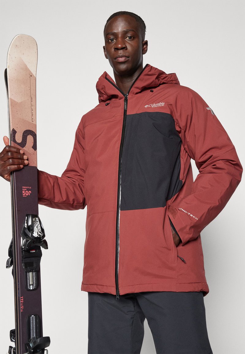 Giacca da sci Columbia in bordeaux e nero, con zip frontale, cappuccio e polsini regolabili, abbinata a sci e attacchi.