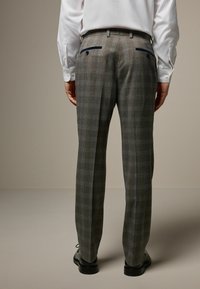Karierte Hose in Grau mit blauen Akzenten. Slim Fit, zwei Gesäßtaschen mit Knopfverschluss. Getragen mit einem weißen Hemd und schwarzen Schuhen.