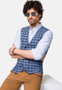 Niebieska kamizelka w kratę na jasnoniebieskiej koszuli z małymi motywami żeglarskimi. Beżowe spodnie i okrągłe okulary przeciwsłoneczne dopełniają strój. Casualowy ubiór.