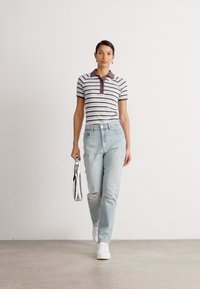 Gestreepte polo met een bruine kraag, lichtblauwe distressed jeans en witte sneakers. Houdt een glanzende zilveren tas vast. Minimalistische achtergrond.