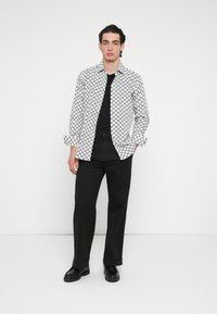 Fehér ing fekete geometriai mintával, fekete póló alatta, fekete nadrág és fekete csizma. A modell sima háttér előtt áll.