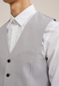 Ljusgrå väst av slät tyg. Har en bröstficka och två bruna knappar. Bärs över en vit skjorta med spetsig krage.