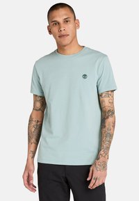 Ljusblå bomulls t-shirt med rund hals och korta ärmar. Den har en liten grön logotyp på bröstet och en mjuk, slät textur.