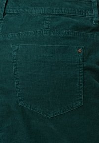 Tessuto in corda color teal con righe verticali, caratterizzato da una tasca a toppa con contorno cucito e un dettaglio in rivetto metallico marrone.