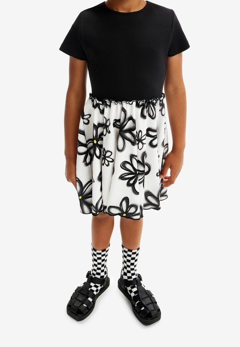 Enfant portant une robe noire à manches courtes avec une jupe blanche ornée de grandes fleurs noires et des chaussettes à carreaux avec des sandales noires.