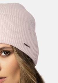 Bonnet en maille côtelée rose pâle avec une légère retombée ; présente un accent logo métallique sur le côté. Gros plan sur le chapeau et l'œil.
