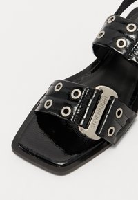 Black patent leather sandal with a square toe, featuring two wide straps adorned with silver hardware and eyelet details.