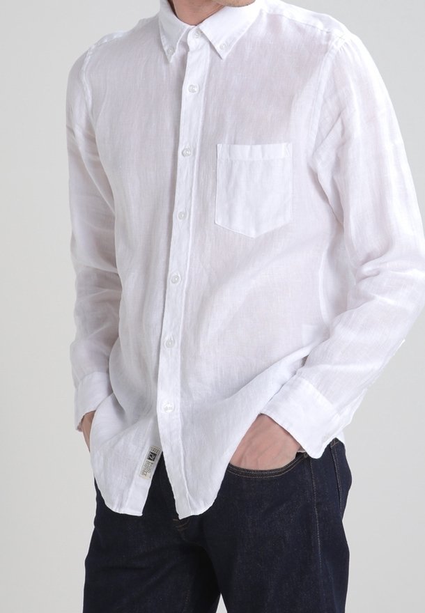 Chemise blanche en lin à manches longues avec col à boutons, poche poitrine unique, tissu légèrement texturé et coupe décontractée. Portée sur un jean foncé.