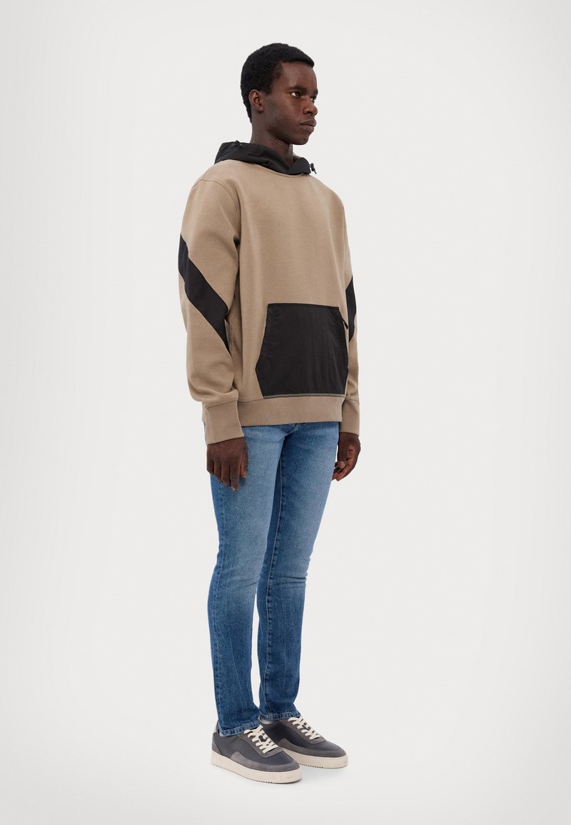 Felpa con cappuccio beige con dettagli neri e tasca frontale, abbinata a jeans slim-fit blu e sneakers grigie. Tessuto morbido, design casual.