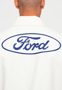 White garment featuring a large blue embroidered "Ford" logo within an oval, showcasing a classic design with a smooth texture.
