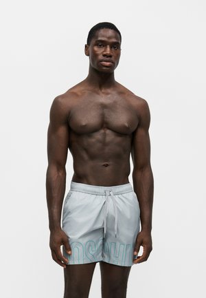 Homme musclé debout portant un short de bain gris clair avec un cordon et des grandes lettres bleues partielles.