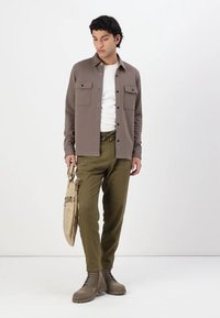 Casaco de camisa castanho com fecho de botão, dois bolsos no peito, t-shirt branca por baixo, calças verdes, bolsa bege e botas castanhas.