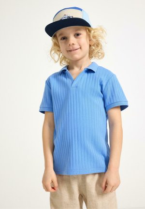 Vaaleatukkainen lapsi, joka pukeutunut siniseen, ribilliseen lyhythihaiseen poolopaitaan ja beigen värisiin housuihin, hymyilee ja käyttää sinis-beigeä lippalakki, jossa on laivakuvio.