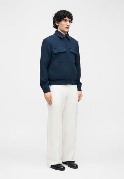 Muž v tmavomodrej bundy na zips s vreckami so záklopkou, bielych širokých nohaviciach a čiernych topánkach stojí na jednoduchom bielom pozadí.