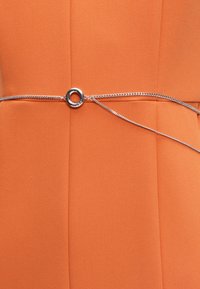 Ceinture fine en chaîne argentée avec une boucle circulaire portée sur un vêtement en tissu orange.