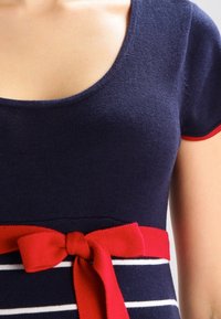 Kvinna som bär en marinblå klänning med korta ärmar och röd kant på ärmarna samt ett rött bälte med rosett ovanför vita horisontella ränder.