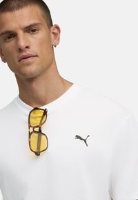 Bílý tričko s malým černým logem Puma, doplněné o brýle v želvovinovém designu se žlutými čočkami, umístěné na tričku.